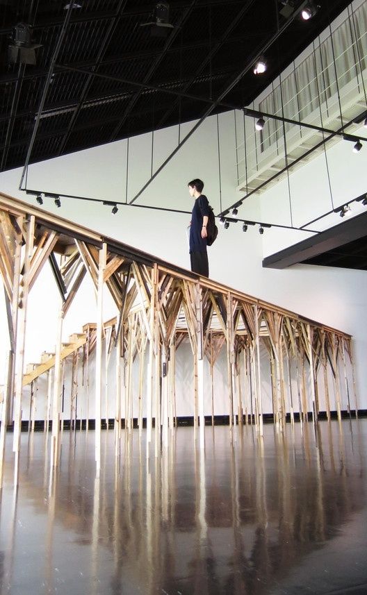 Person standing atop a large timber framework installation in a gallery